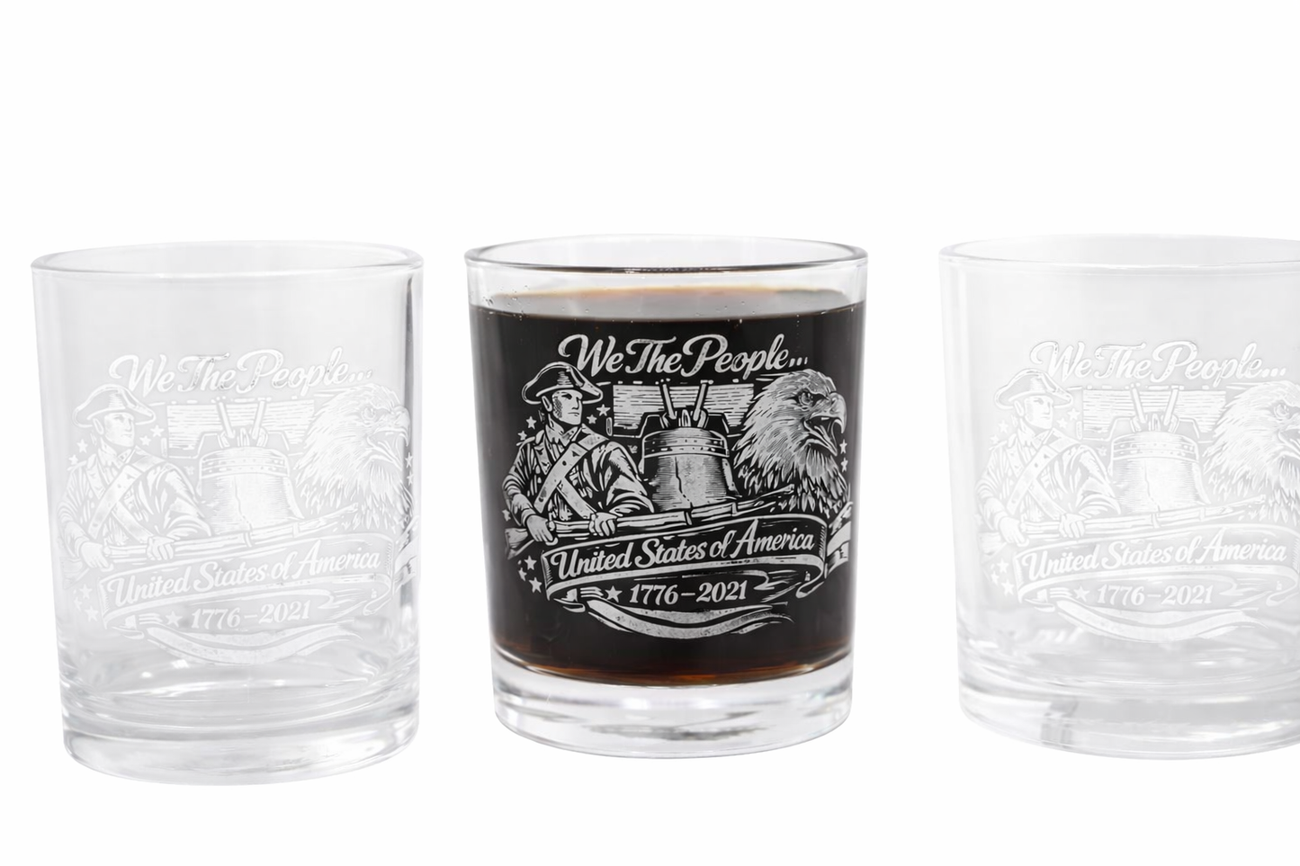Three clear whiskey glasses with engraved patriotic artwork featuring a colonial soldier, Liberty Bell, and bald eagle. The center glass is filled with dark whiskey, while the two on each side are empty