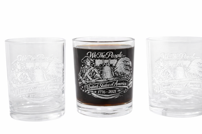 Three clear whiskey glasses with engraved patriotic artwork featuring a colonial soldier, Liberty Bell, and bald eagle. The center glass is filled with dark whiskey, while the two on each side are empty