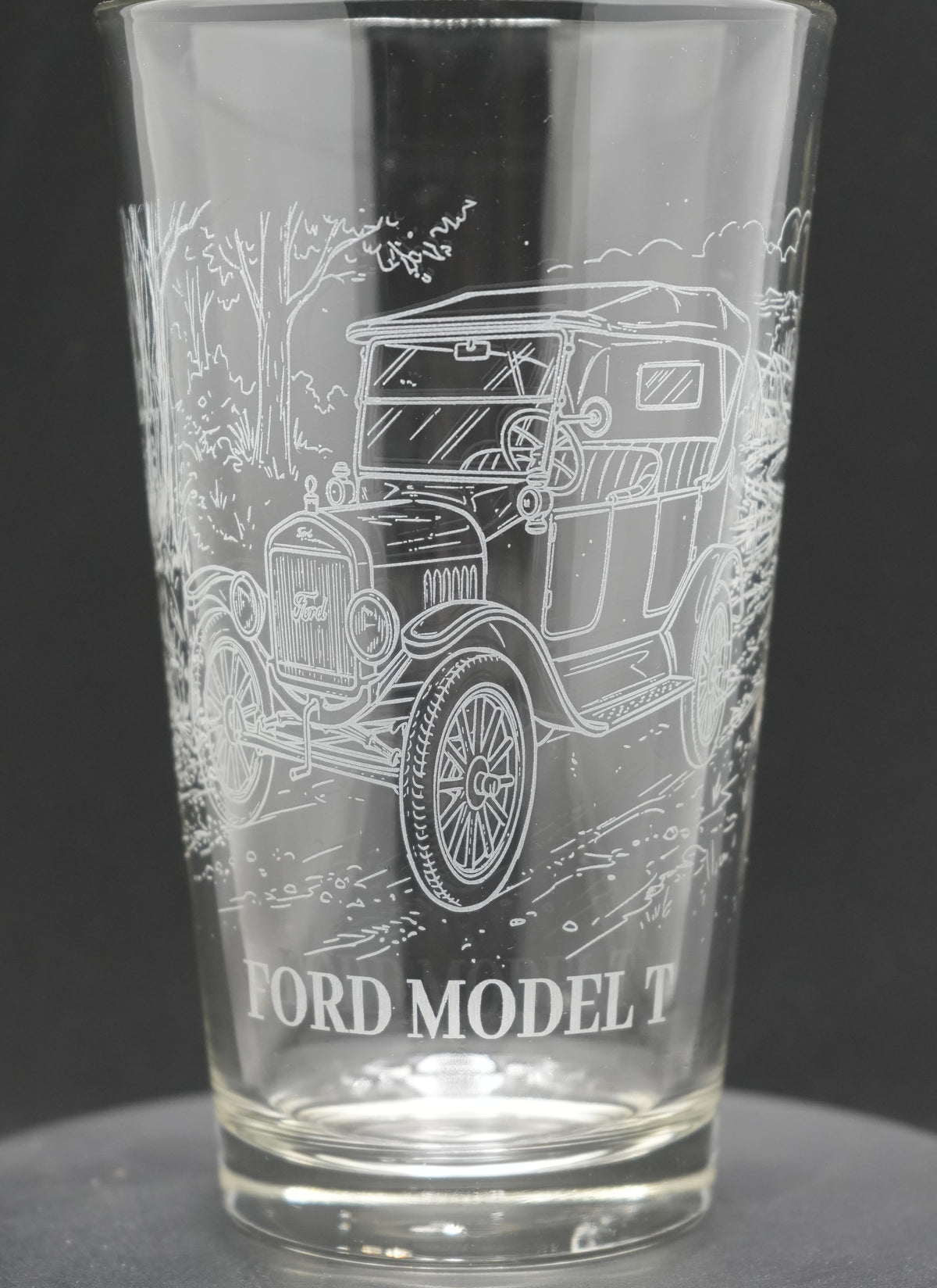 A clear glass pint tumbler featuring an etched white line drawing of a vintage Ford Model T car, with 'FORD MODEL T' text printed near the base, set against a dark gray background.