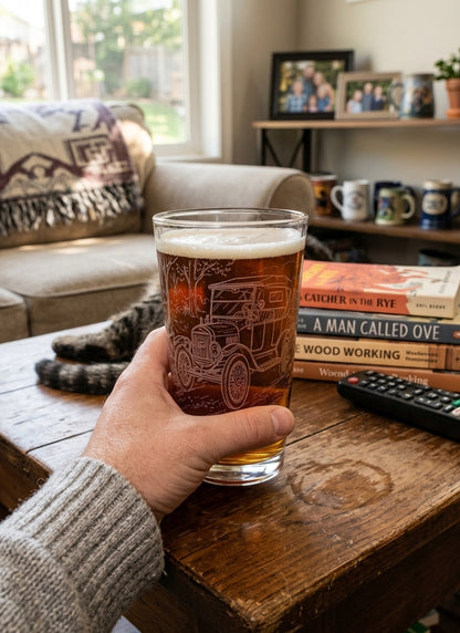 Hand holding a 16 oz Ford Model T engraved beer pint glass filled with amber beer, in a living room setting.