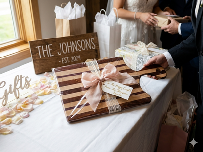 A handcrafted striped wood charcuterie board wrapped in a pink ribbon and lace bow with a gift tag, displayed on a wedding reception gift table with a bride and groom in the background.
