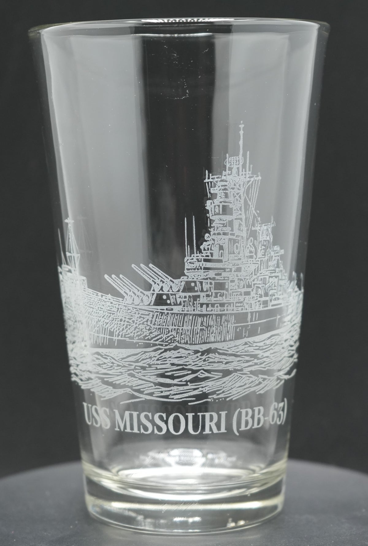 A clear glass pint glass featuring an etched illustration of the USS Missouri (BB-61) warship and its name, shown against a dark gray background.